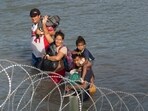 Migrants wave as they walk near concertina wire in the water along the Rio Grande border with Mexico in Eagle Pass, Texas, on July 16. Migrants wave as they walk near concertina wire in the water along the Rio Grande border with Mexico in Eagle Pass, Texas, on July 16.