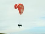 The image shows an artist playing the piano while flying with the help of a parachute. The image shows an artist playing the piano while flying with the help of a parachute.