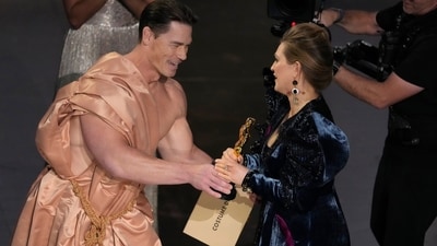 John Cena, left, presents the award for best costume design to Holly Waddington for Poor Things. As the nomination were announced, a team helped him wear a silk robe so he could use his hands to open the envelope and make the announcement. (Chris Pizzello/Invision/AP)