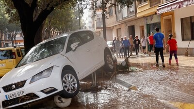 A damaged car sits elevated as people walk along a mud-covered street after torrential rains caused flooding in La Alcudia, Valencia. (REUTERS)
