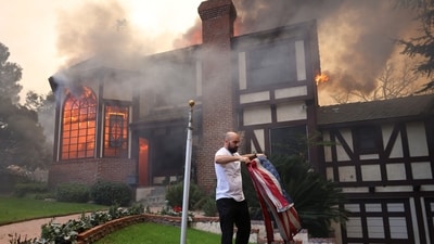 A person folds a flag after lowering it from the flagpole outside his burning cousin's house as powerful winds fueling devastating wildfires in the Los Angeles area force people to evacuate, at the Eaton Fire in Altadena, California, (REUTERS)