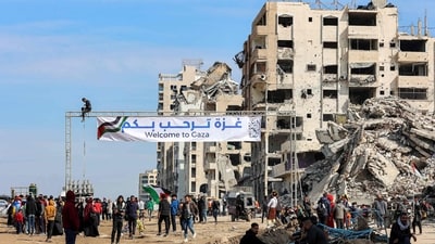 People gather by a banner welcoming people near the rubble of a collapsed building along Gaza's coastal al-Rashid Street for people to cross from the Israeli-blocked Netzarim corridor from the southern Gaza Strip into Gaza City on January 26, 2025. Israel said on January 25 it would block the return of displaced Palestinians to their homes in northern Gaza until Arbel Yehud, one of the hostages taken captive during the October 7, 2023 attacks, is released. Hamas sources said that Yehud was "alive and in good health", and would be "released as part of the third swap set for next Saturday", on February 1. (Photo by Omar AL-QATTAA / AFP) (AFP) People gather by a banner welcoming people near the rubble of a collapsed building along Gaza's coastal al-Rashid Street for people to cross from the Israeli-blocked Netzarim corridor from the southern Gaza Strip into Gaza City on January 26, 2025. Israel said on January 25 it would block the return of displaced Palestinians to their homes in northern Gaza until Arbel Yehud, one of the hostages taken captive during the October 7, 2023 attacks, is released. Hamas sources said that Yehud was "alive and in good health", and would be "released as part of the third swap set for next Saturday", on February 1. (Photo by Omar AL-QATTAA / AFP) (AFP)