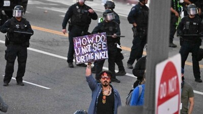 A demonstrator holds a sigh reading "How Do U Not See Yur the Nazi" while police officers in riot gear move to clear the street as curfew comes into effect after several days of protests Los Angeles. (AFP) A demonstrator holds a sigh reading "How Do U Not See Yur the Nazi" while police officers in riot gear move to clear the street as curfew comes into effect after several days of protests Los Angeles. (AFP)