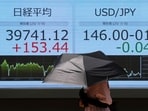 A woman holding a sunshade walks in front of a screen displaying Japan's Nikkei share average and exchange rate between Japanese yen and US dollar outside a brokerage in Tokyo