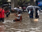 A man sits on a chair as people wade through a waterlogged road amid rainfall, in Mumbai, Tuesday