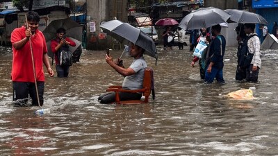 A man sits on a chair as people wade through a waterlogged road amid rainfall, in Mumbai, Tuesday (PTI) A man sits on a chair as people wade through a waterlogged road amid rainfall, in Mumbai, Tuesday (PTI)