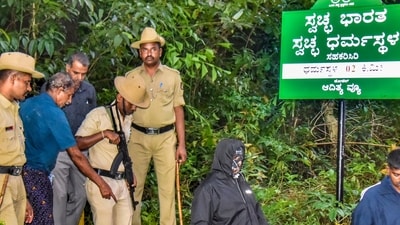 Security personnel with the whistleblower, who claims to have been coerced into burying and cremating bodies of women and minors, near a site of an alleged burial related to the Dharmasthala mass burial case, at Dharmasthala, in Dakshina Kannada district, (PTI) Security personnel with the whistleblower, who claims to have been coerced into burying and cremating bodies of women and minors, near a site of an alleged burial related to the Dharmasthala mass burial case, at Dharmasthala, in Dakshina Kannada district, (PTI)