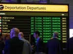 Participants stand in front of a build-up 'Deportation Departures board' before British Reform party leader Nigel Farage addresses journalists during a press conference in a hangar at Oxford Airport in Kidlington, England, Aug. 26
