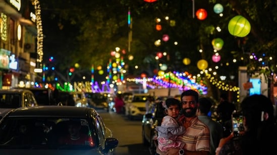 New Delhi: A man holds a child while a family member takes photographs on a street illuminated with lights on the eve of the �Diwali� festival, in New Delhi, Sunday, Oct. 19, 2025. (PTI Photo/Salman Ali) (PTI10_19_2025_000297B)(PTI)