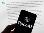 The OpenAI logo is displayed on a mobile phone in front of a computer screen with output from ChatGPT.