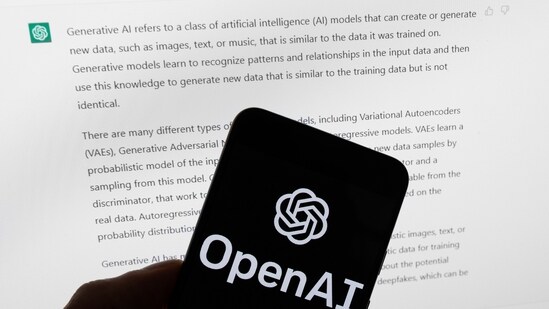 The OpenAI logo is displayed on a mobile phone in front of a computer screen with output from ChatGPT. (AP)