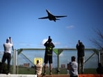 Plane spotters stand at a perimeter fence to see and photograph US Air Force (USAF) B-1 Lancer bomber jets taking off from a base