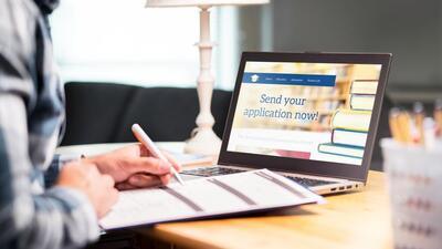 Young man writing college or university application form with pen and paper. Student applying or filling document. Scholarship, grant or admission. School website in laptop. (Getty Images/iStockphoto)