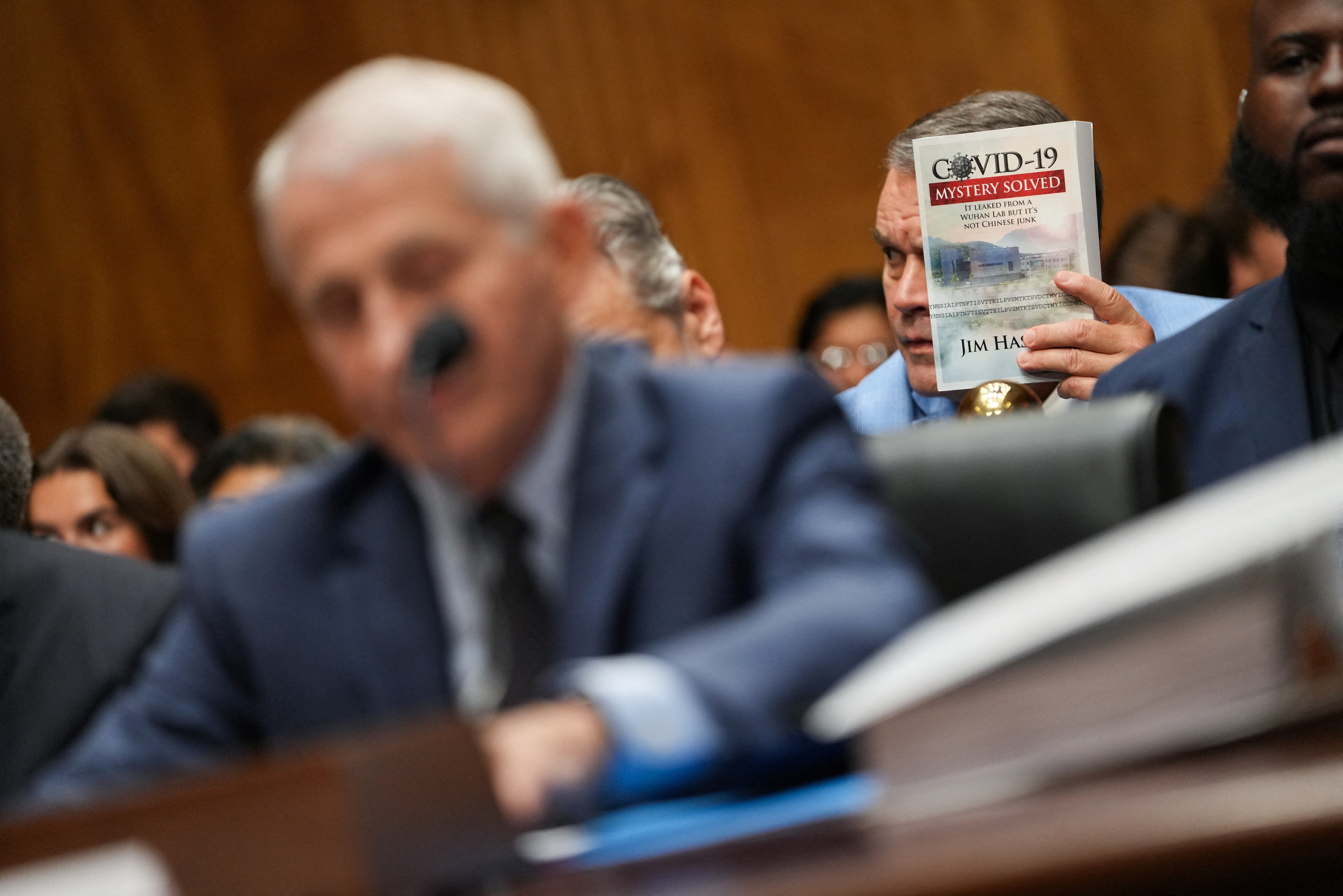 A man holds a book 'COVID-19: Mystery Solved: It leaked from a Wuhan lab but it's not Chinese junk' behind Anthony Fauci, the former director of the US National Institute of Allergy and Infectious Diseases (NIAID), on Capitol Hill in Washington, DC. (Reuters Photo)