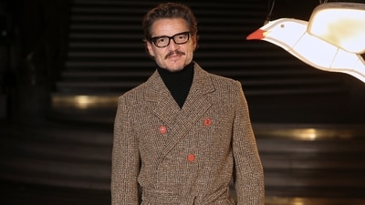 Pedro Pascal poses for the press upon arrival at the Chanel showcase. He wore a wool trench coat blazer styled with a black turtleneck sweater, nerdy glasses, and pants.&nbsp; (AP Photo/Thomas Padilla)