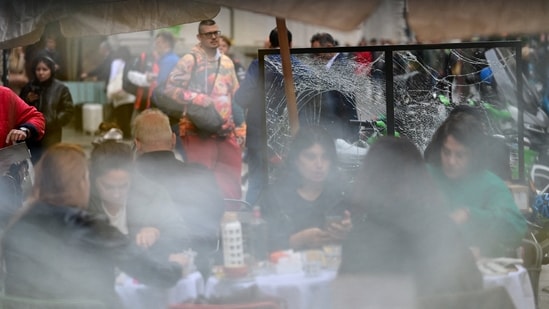 People sit at an outdoor cafe near a shattered glass panel following weekend clashes between police and protesters in Milan, Italy, October 19, 2026.